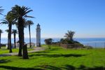 A tall white-walled cylindrical lighthouse sits at the end of a road that curves through a grassy field. Palm trees (and one stump) flank the road, while several squarish buildings cluster near its base. Beyond, you can see the ocean and a hazy blue sky.