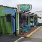 Small building painted green next to a sidewalk lined with palm trees. Dried palm fronds cover an awning, and two small round tables, each with a pair of chairs, flank an open glass door.