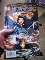 A comic book, held in my left hand. It's New Spring #8, published by Dynamite Comics. It features a white-looking woman with black hair in a blue dress with a blue cloak and a jeweled forehead ornament. Her hair and cloak are billowing in wind. Her fingertips rest lightly on the head of a man kneeling in front of her, facing the reader, his head lowered slightly and his eyes closed. Below the comic book a tiled floor and the bottom of a bookshelf are visible, because I had to get photographic evidence that this thing was actually real as soon as I picked it up, before I even paid for it and left the shop.