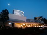 A wide, low building with an overhang lit up by *lots* of lights, against a dark blue sky. Two taller glass-walled office buildings are visible behind it, as is the bright splotch of the moon. Silhouetts of trees and a spiky hedge frame the sides and bottom of the frame. The words SOUTH COAST can be seen running along the overhang.