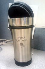 A tall, brushed-metal travel mug sitting on a table, its black plastic/rubber lid propped open. There's no handle, and the slogan 'Venti Schmenti' appears prominently on the side. Below it, barely legible, are the Diedrich Coffee logo and the phrase 'Not so big.'