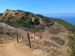A winding dirt trail with posts and grab ropes on one side leads down a hillside into a narrow valley. A ridge of jagged rocks rises beyond it. A trail is visible curving around the ridge, while a gentle hillside slopes down toward the ocean. The nearby hill is mostly dry grass, but green trees line the valley and brush